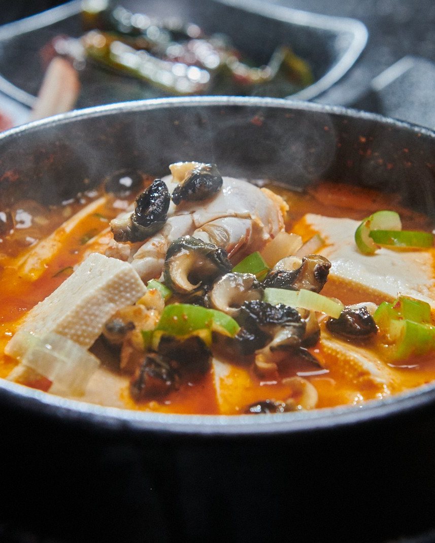 석화한우암소생구이 제주 보말 한우 뽀글된장 — 검은 냄비에 자박하게 끓인 주황빛 된장찌개. 두부와 검은 보말, 송송 썬 풋고추와 대파가 보인다.