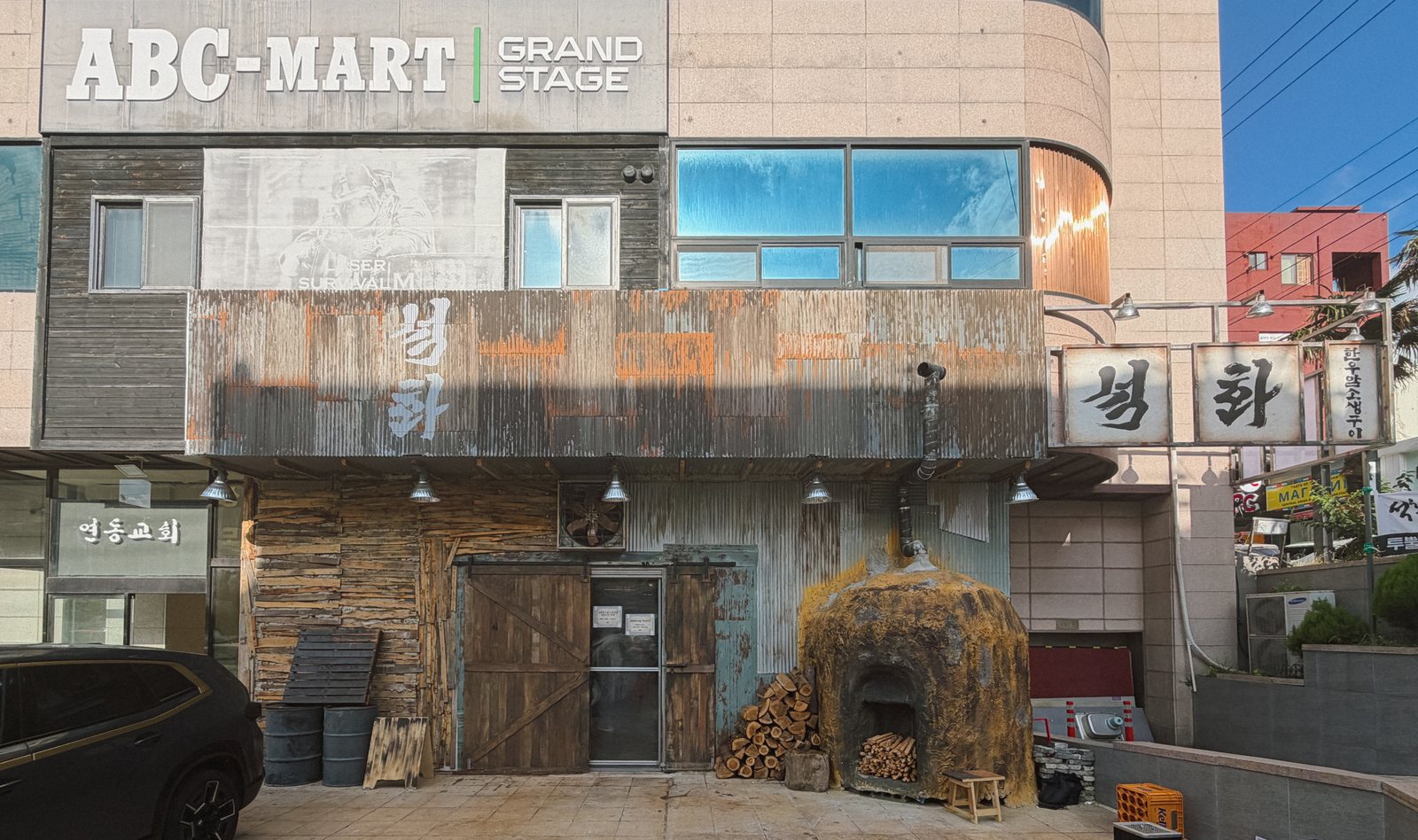 석화한우암소생구이 외관 — 맑은 낮에 본 상가 건물. 위층 벽에 ‘ABC-MART GRAND STAGE’ 간판이 걸려 있고, 1층 매장은 녹슨 골함석과 낡은 판자 외벽에 흰 붓글씨 ‘석화’, 오른쪽 모서리에 ‘석화’ 돌출 간판, 입구 옆 흙 화덕과 장작더미가 보인다. 왼쪽 아래에 주차된 검은 승용차 일부가 있다.