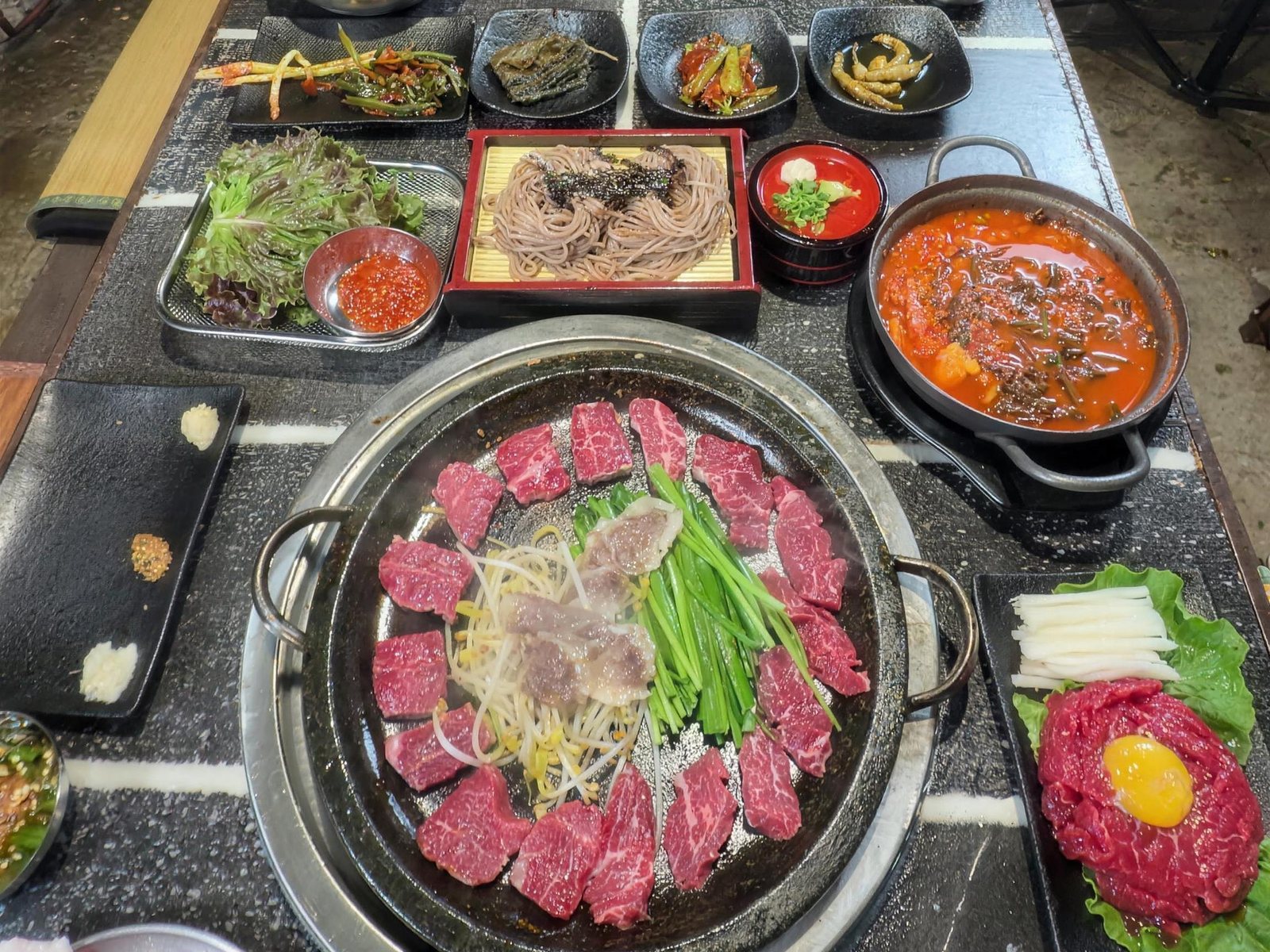 석화한우암소생구이 상차림 — 테이블에 차린 한 상을 위에서 본 모습. 앞쪽 둥근 돌판에 한우를 둥글게 두르고 가운데 숙주와 부추를 올렸고, 오른쪽 아래에 노른자를 올린 육회, 가운데 위쪽에 판소바, 오른쪽에 빨간 찌개 냄비, 왼쪽에 쌈채소와 반찬 접시들이 놓여 있다.