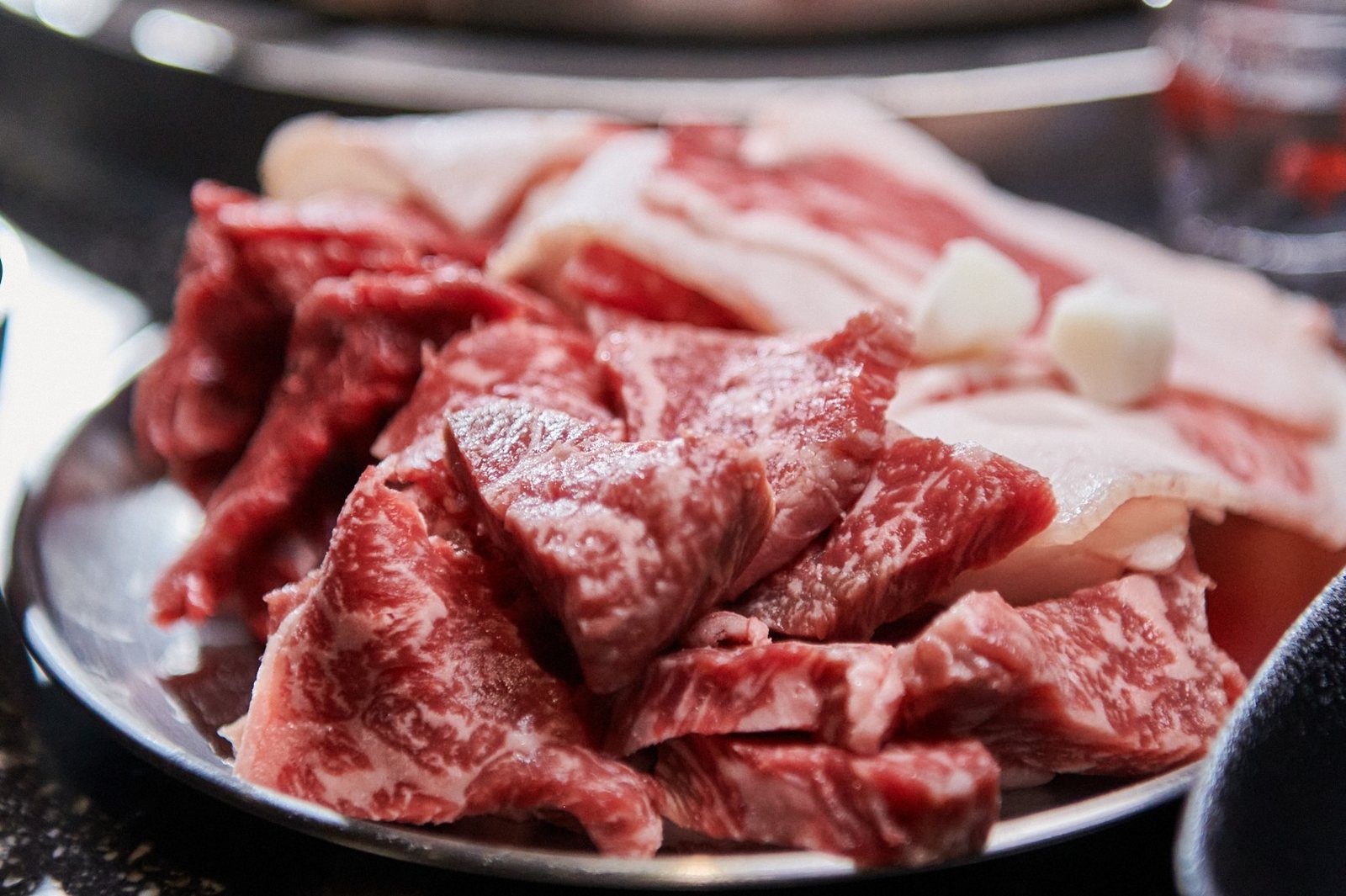 석화한우암소생구이 석화한우 암소 생구이 소(450g) — 스테인리스 접시에 담긴 생 한우를 가까이 찍은 모습. 마블링이 촘촘한 붉은 덩어리 부위와 흰 지방 줄무늬 슬라이스가 겹쳐 있고, 그 위에 깐 마늘 두 쪽이 올라가 있다.