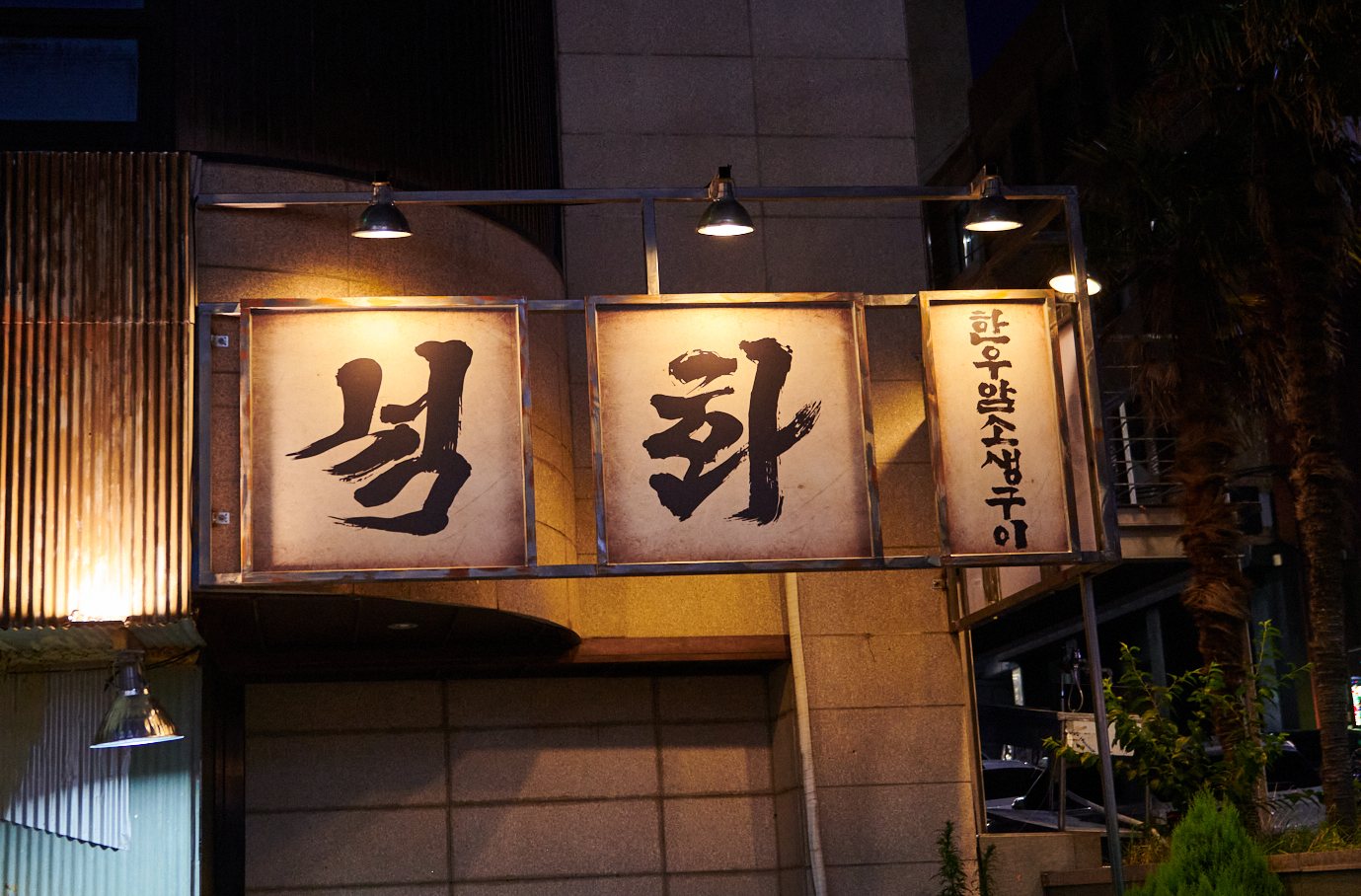 석화한우암소생구이 외관 — 밤에 조명을 받은 돌출 간판. 녹슨 쇠틀 안 흰 판 두 장에 검은 붓글씨 ‘석’·‘화’, 오른쪽 세로 판에 ‘한우암소생구이’가 쓰여 있고 위에 은색 갓 조명이 매달려 있다. 왼쪽에 녹슨 골함석 벽이 조금 보인다.