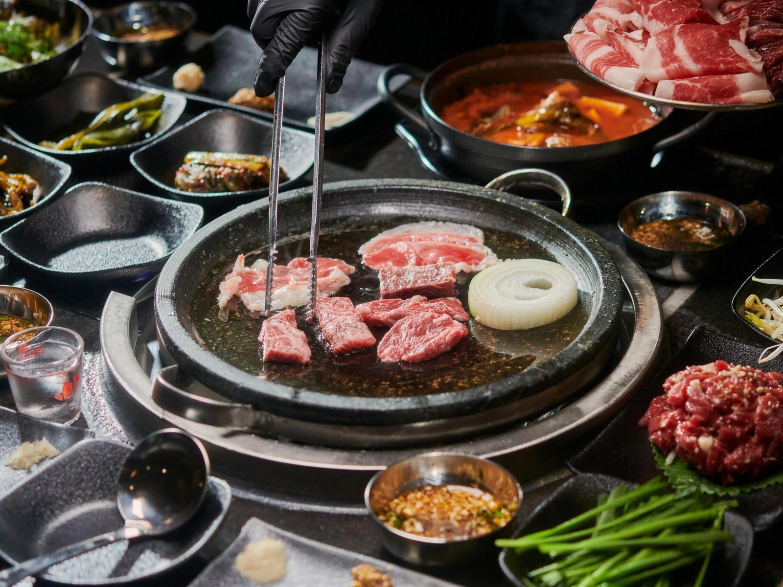 석화한우암소생구이 상차림 — 검은 장갑을 낀 손이 긴 집게로 둥근 돌판 위 한우와 양파 링을 굽는 한 상. 뒤로 빨간 찌개 냄비와 생고기 접시, 왼쪽에 고추장아찌와 김치 접시, 오른쪽 아래에 깻잎에 올린 육회와 부추가 놓여 있다.