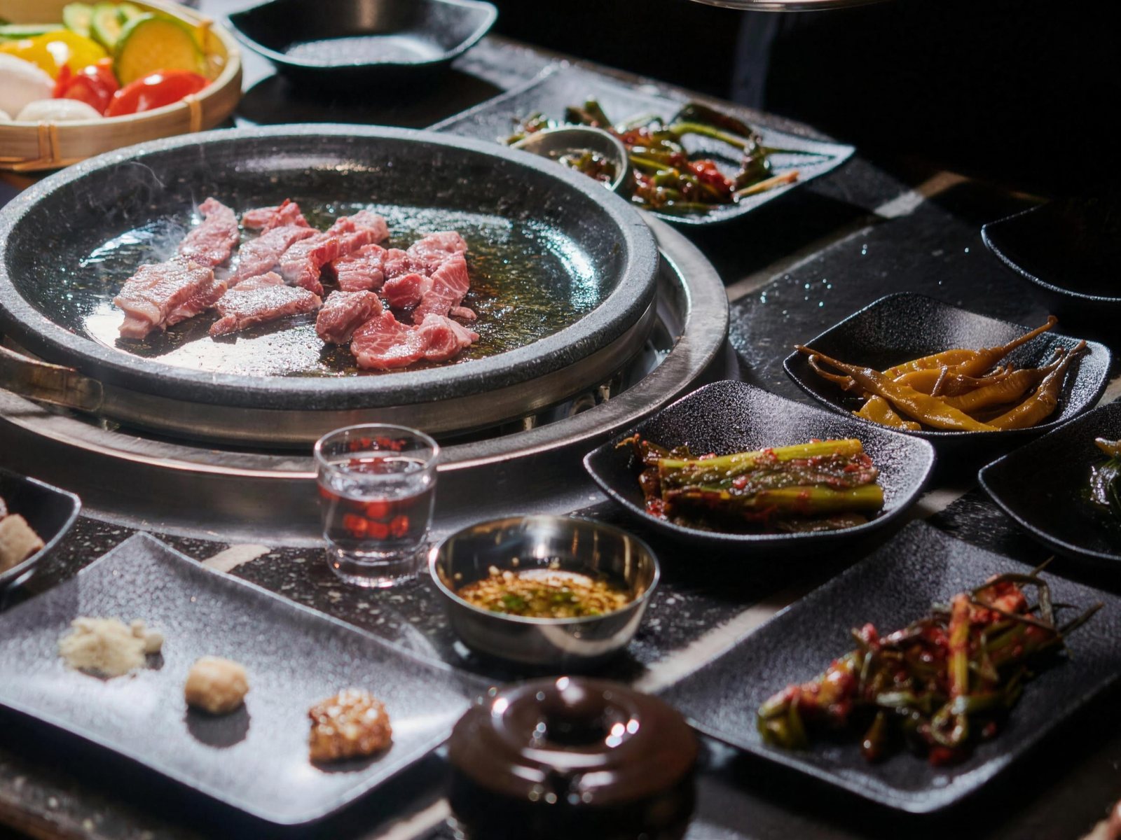 석화한우암소생구이 상차림 — 둥근 돌판에 한우를 올려 굽기 시작한 한 상. 둘레에 고추장아찌와 김치 접시, 마늘·고추를 넣은 간장 소스 그릇, 양념 세 가지를 올린 검은 사각 접시가 놓여 있다.