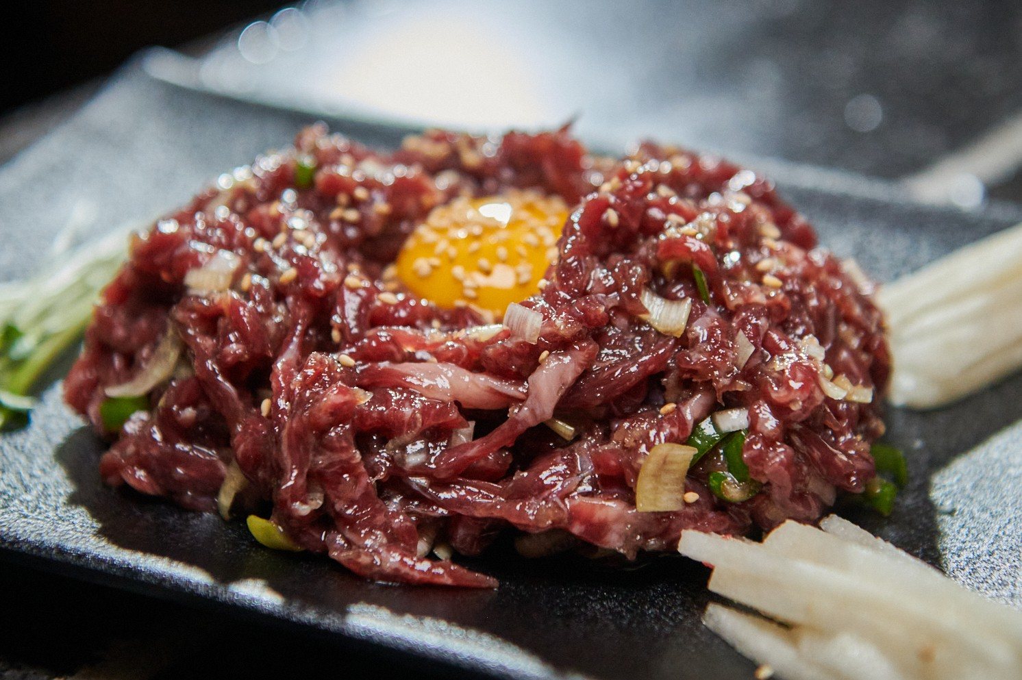 석화한우암소생구이 석화한우 마농육회(180g) — 검은 사각 접시에 수북이 담은 붉은 육회. 노른자와 깨, 송송 썬 파가 올라가 있고 오른쪽 앞에 채 썬 배가 놓여 있다.